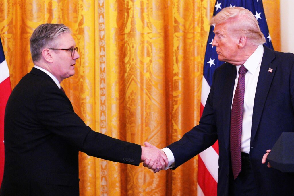 UK and US officials in formal suits shaking hands in front of gold curtains and national flags, symbolizing a diplomatic trade agreement.